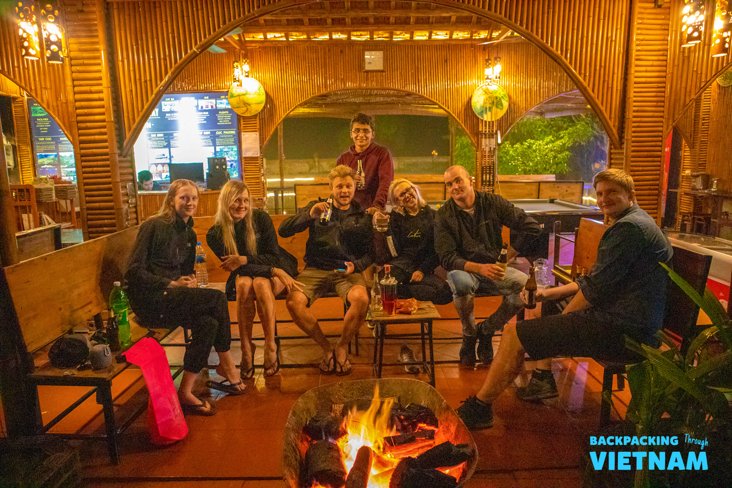 Happy travelers socialize around a warm fire pit, enjoying drinks in a bamboo-decorated setting in Vietnam.