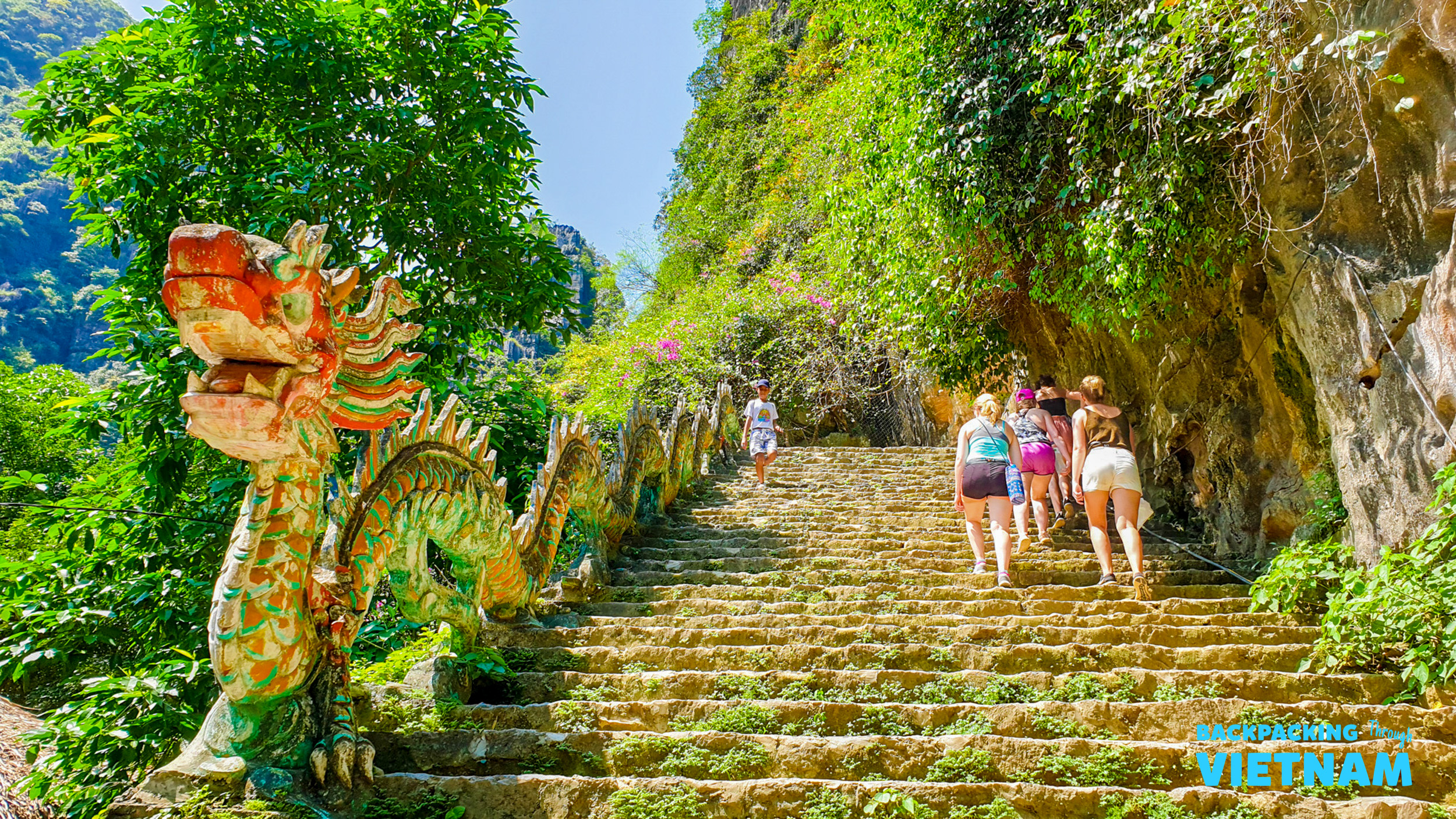 Travelers ascend the ancient stone steps winding past a vibrant dragon sculpture at Hang Mua, Vietnam.