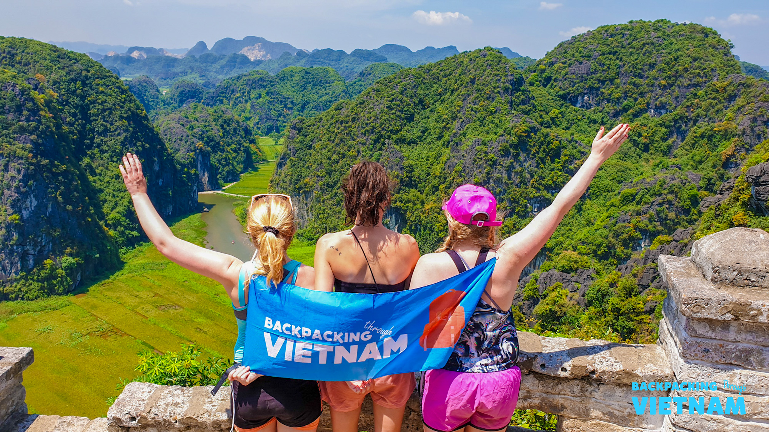 Three joyful women with a "Backpacking Vietnam" flag admire the panoramic view of Tam Coc.