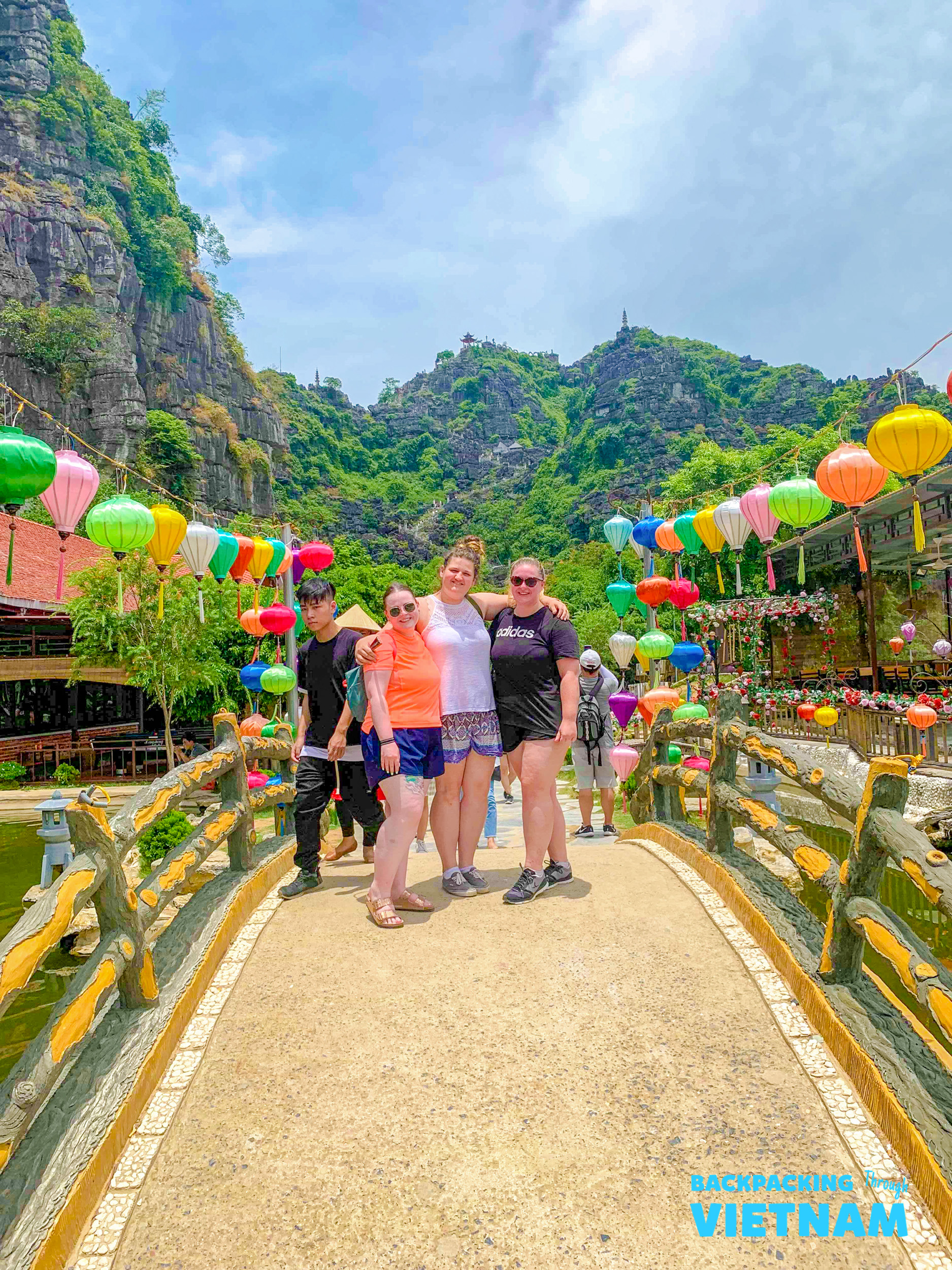 Four happy travelers pose on a colorful bridge with vibrant lanterns and lush mountains in Tam Coc, Vietnam.