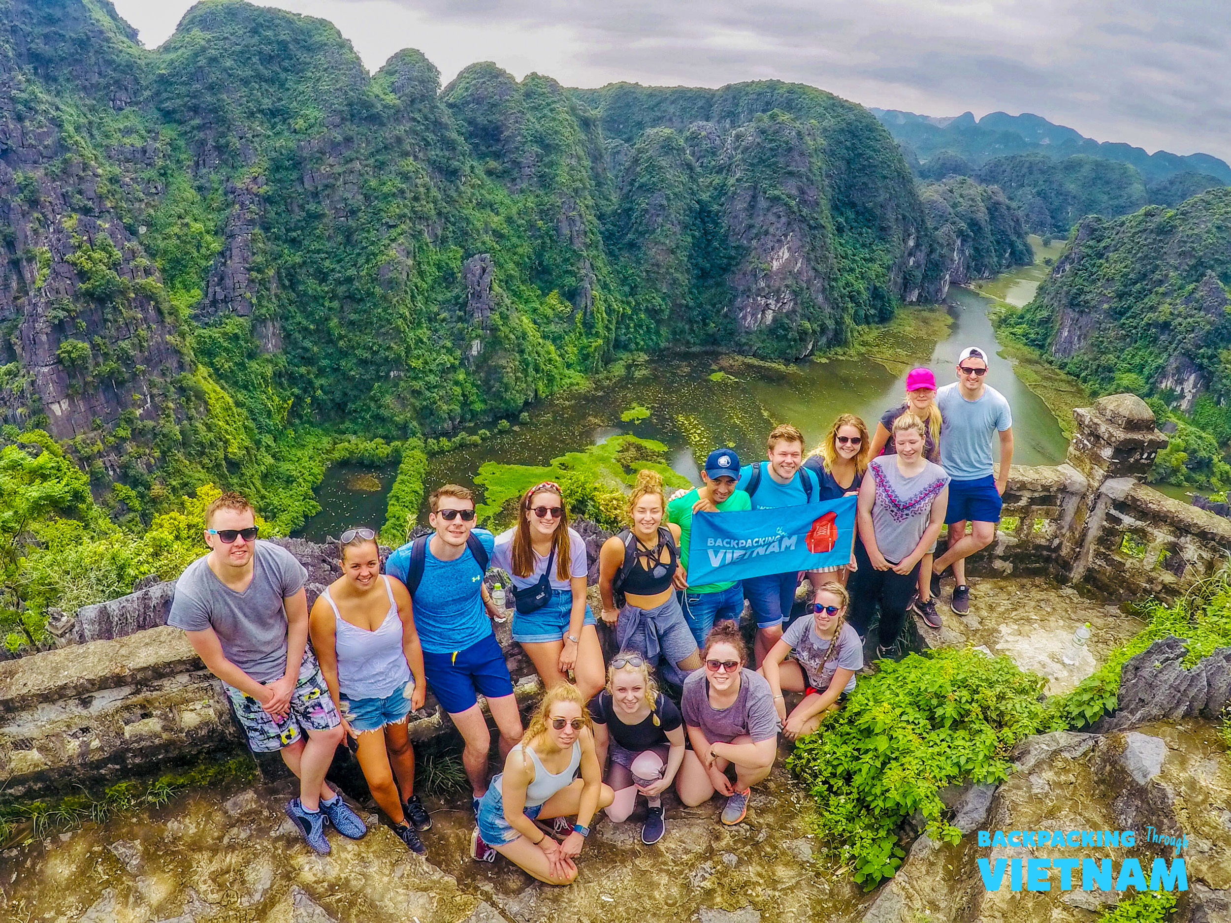 A group of happy backpackers pose at a scenic viewpoint overlooking the lush Tam Coc river landscape.