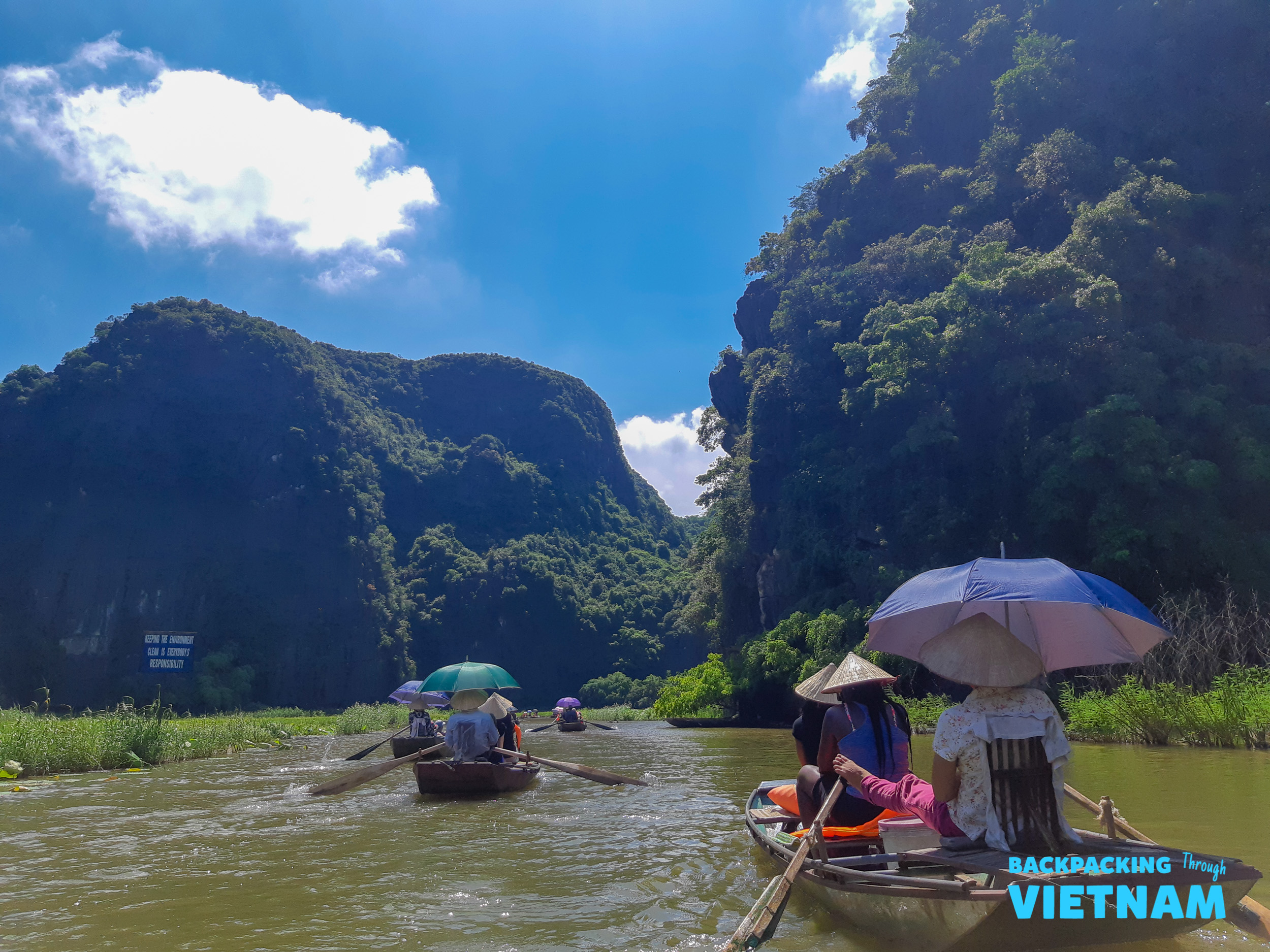 Tourists in traditional boats paddle along a calm river surrounded by towering green karstic mountains in Tam Coc, Vietnam.