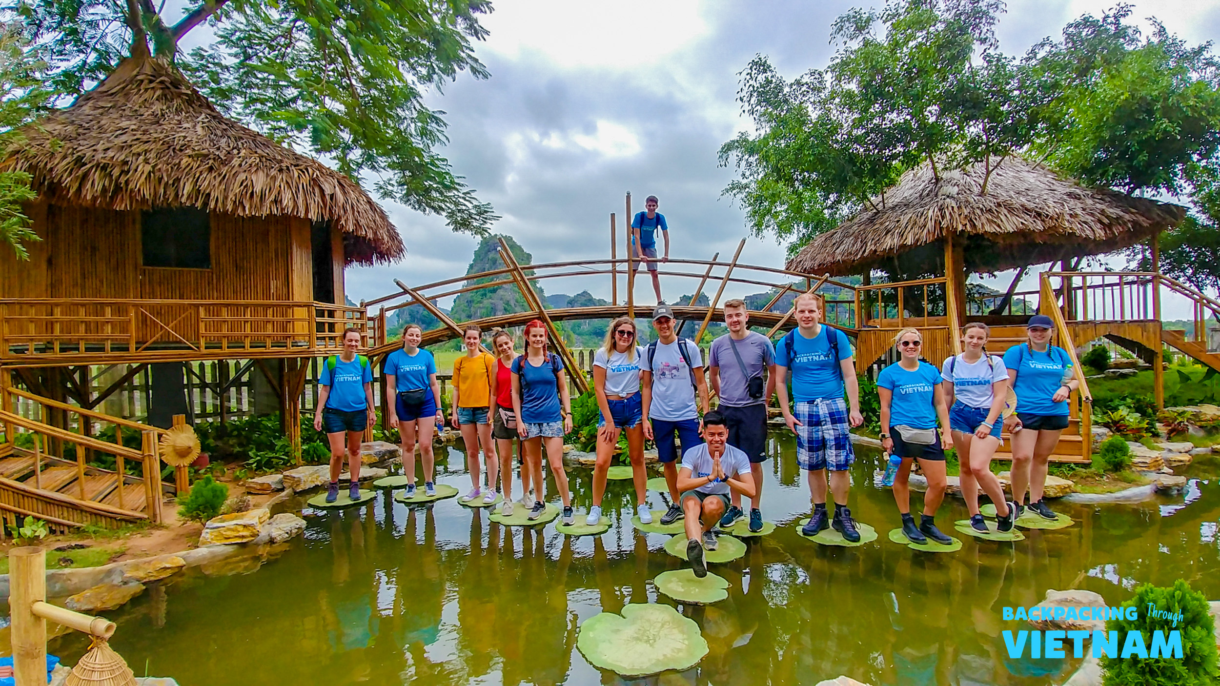 Smiling backpackers pose on lily pads in a pond with traditional huts and karst mountains in Tam Coc.