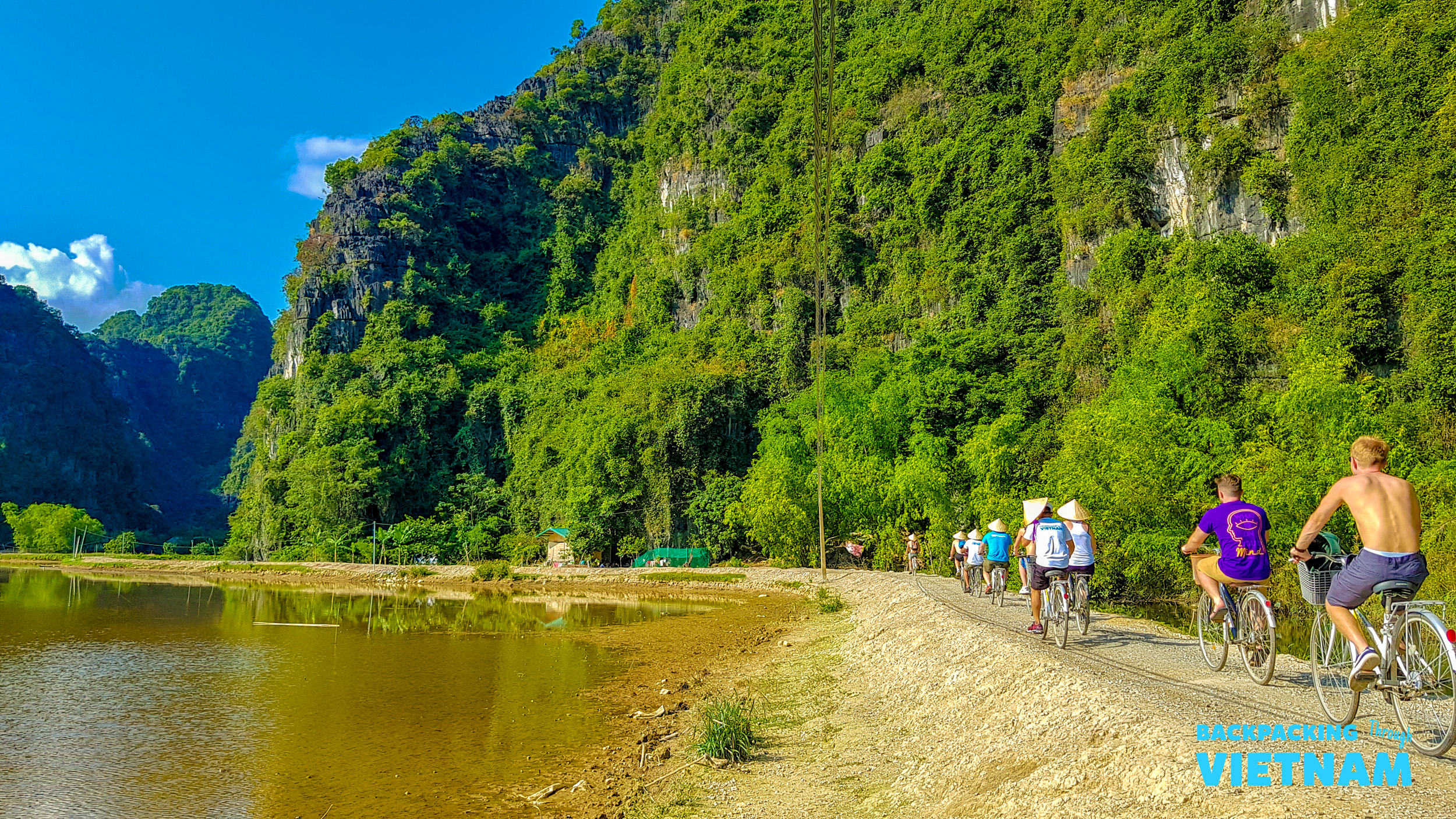 Tourists cycle along a dirt path beside a river with towering green karsts in Tam Coc, Vietnam.