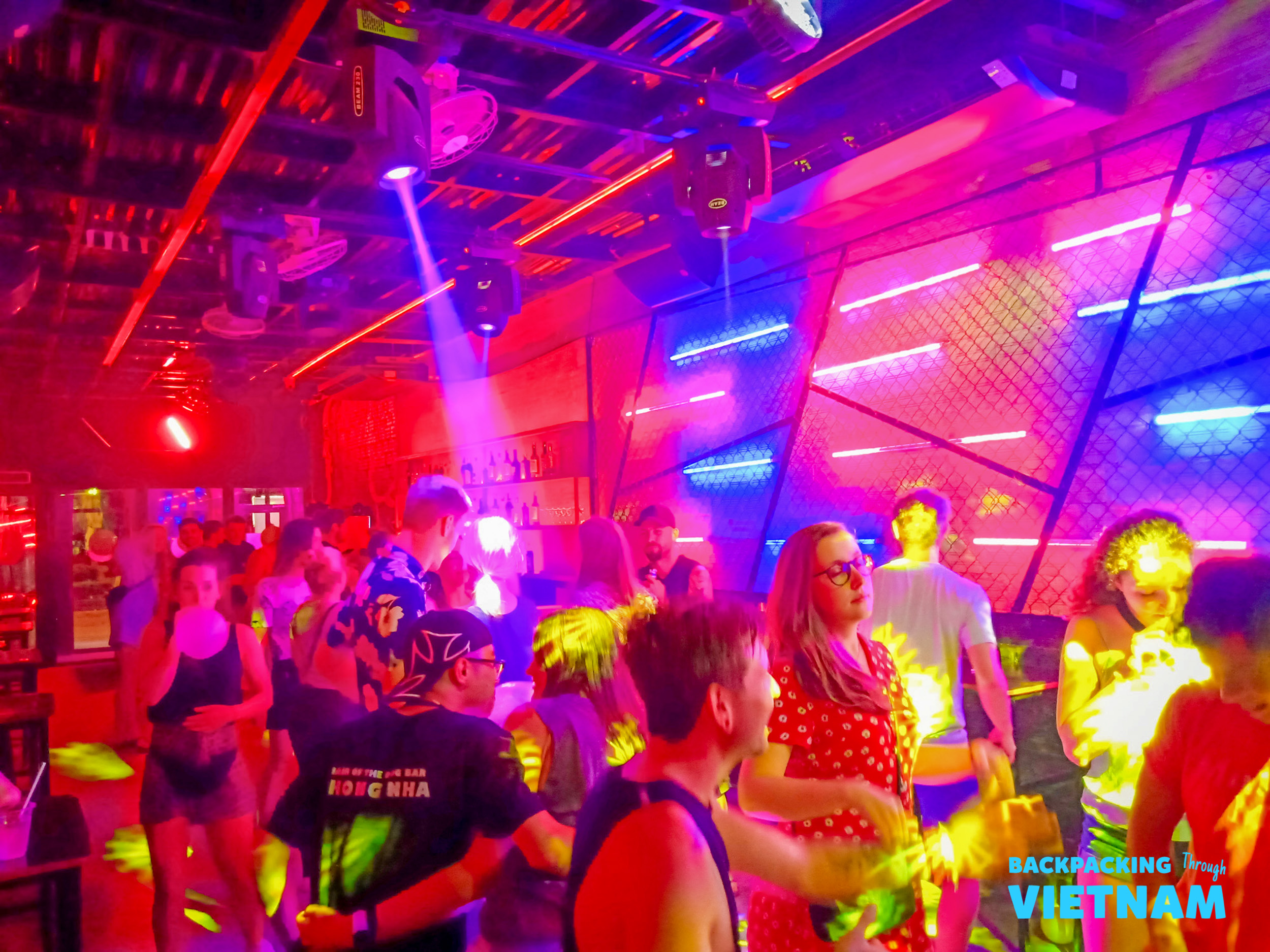 Young travelers dancing and socializing under vibrant red and blue lights in a Phong Nha nightclub.