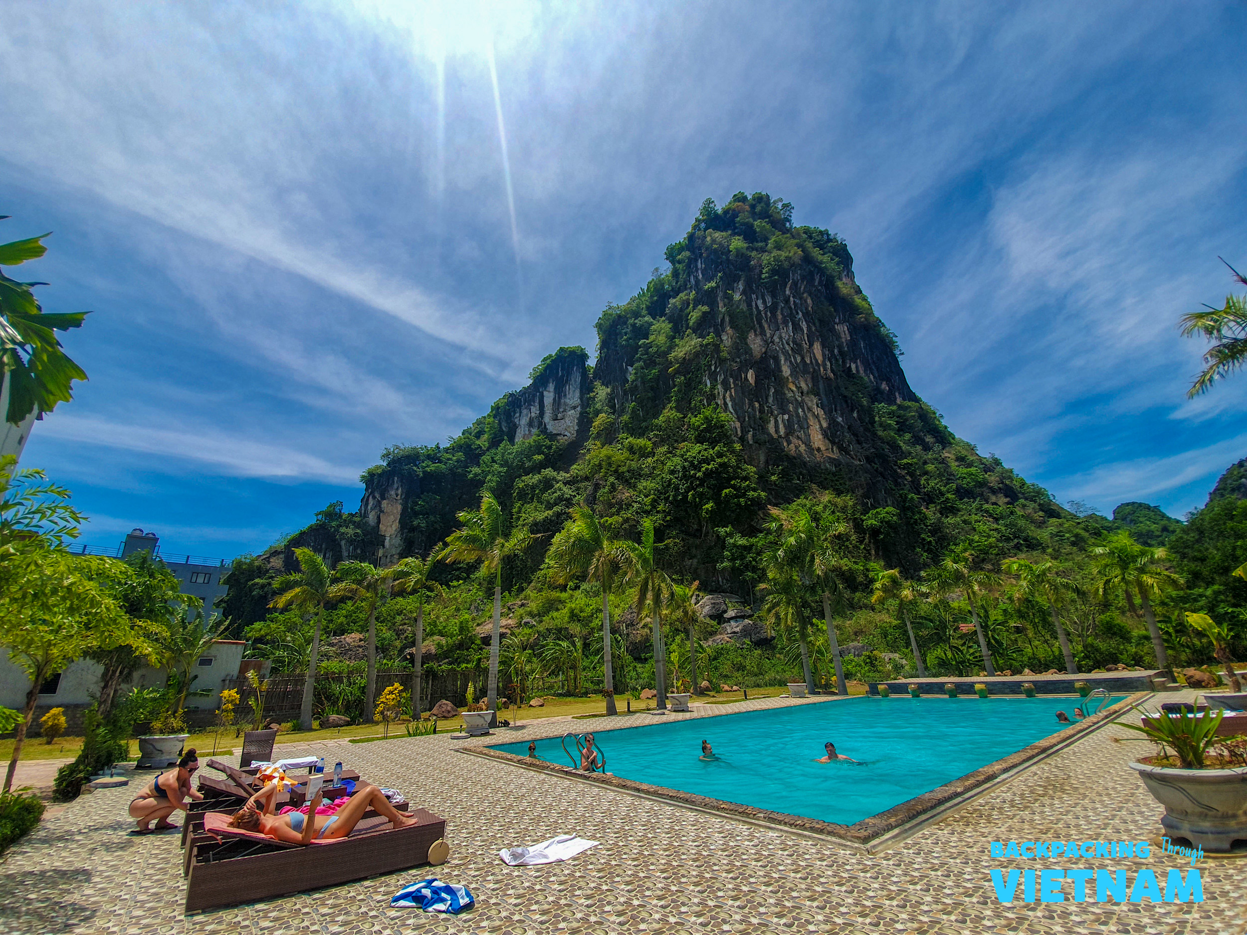 People relax by a bright blue pool with a towering green karst mountain backdrop in Phong Nha, Vietnam.