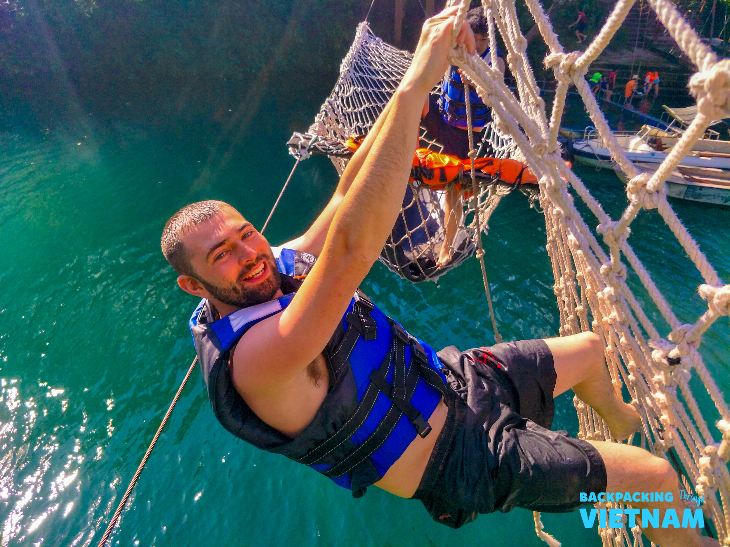 Smiling man in a life vest navigates a rope net obstacle course over water in Phong Nha.