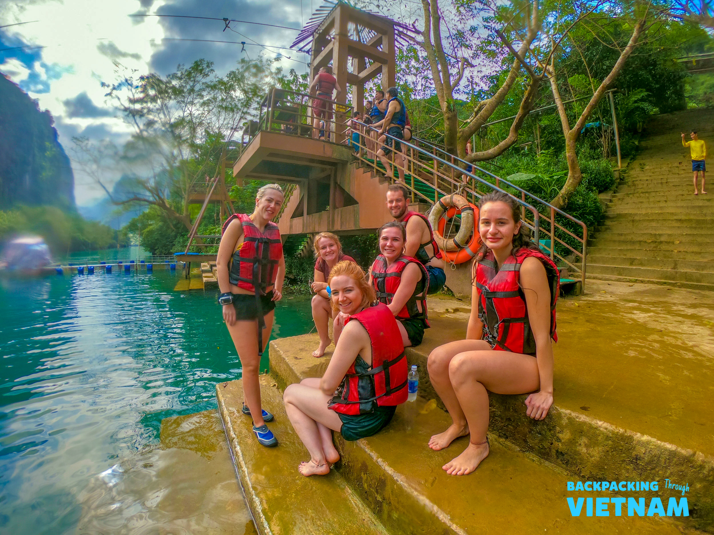 Happy backpackers in red life vests pose by the clear water and zipline platform in Phong Nha.