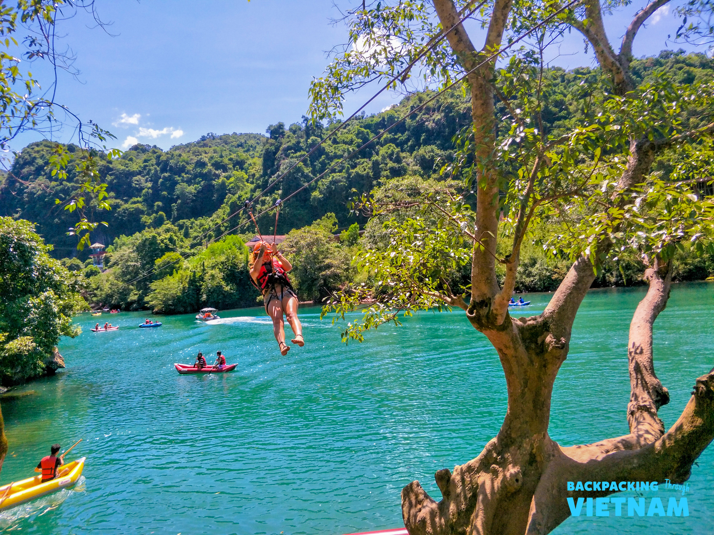 A person ziplines over the clear turquoise river in Phong Nha, with kayakers and green hills.