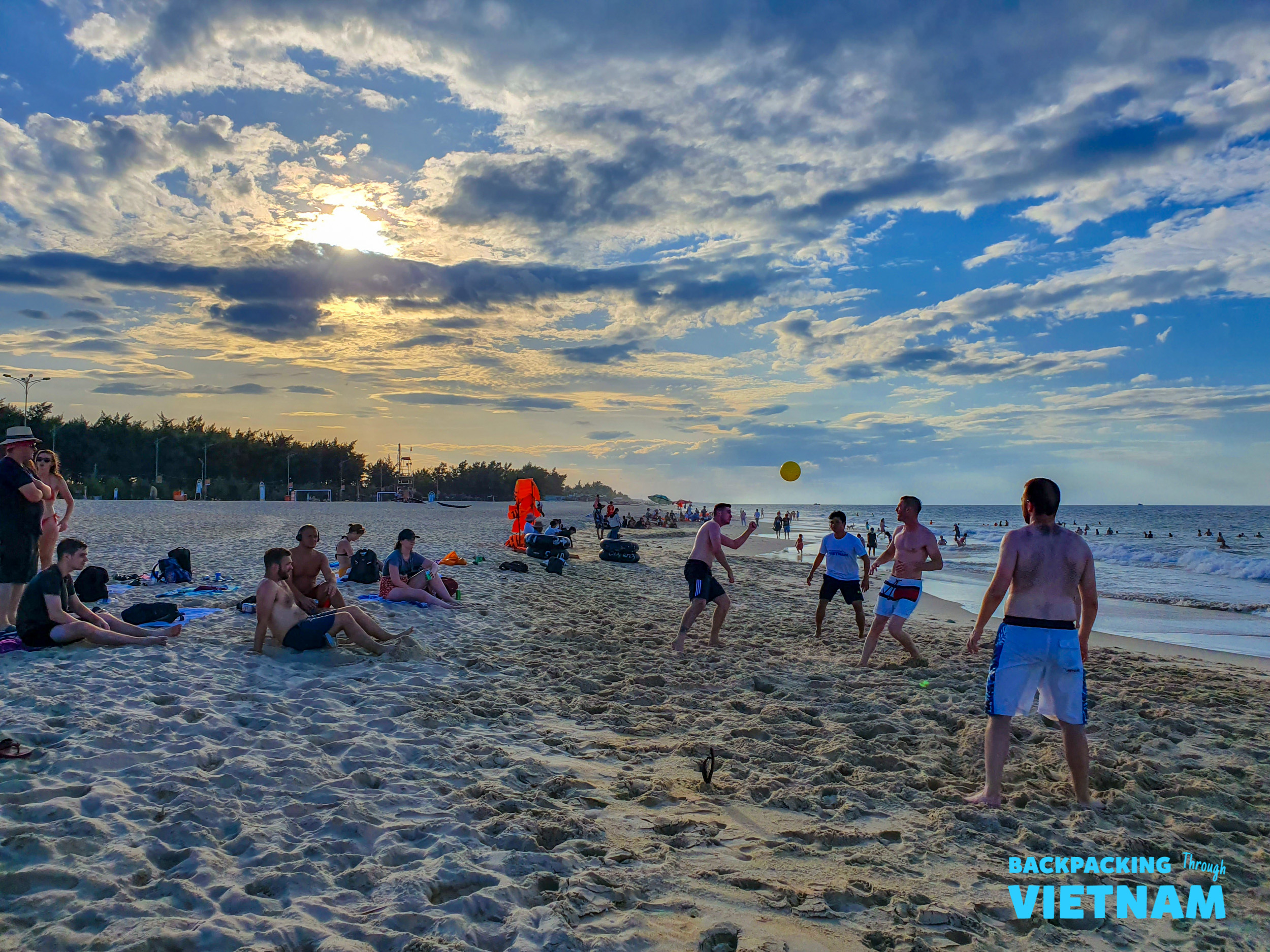 Travelers play volleyball on a sandy beach, enjoying the sunset and ocean views.