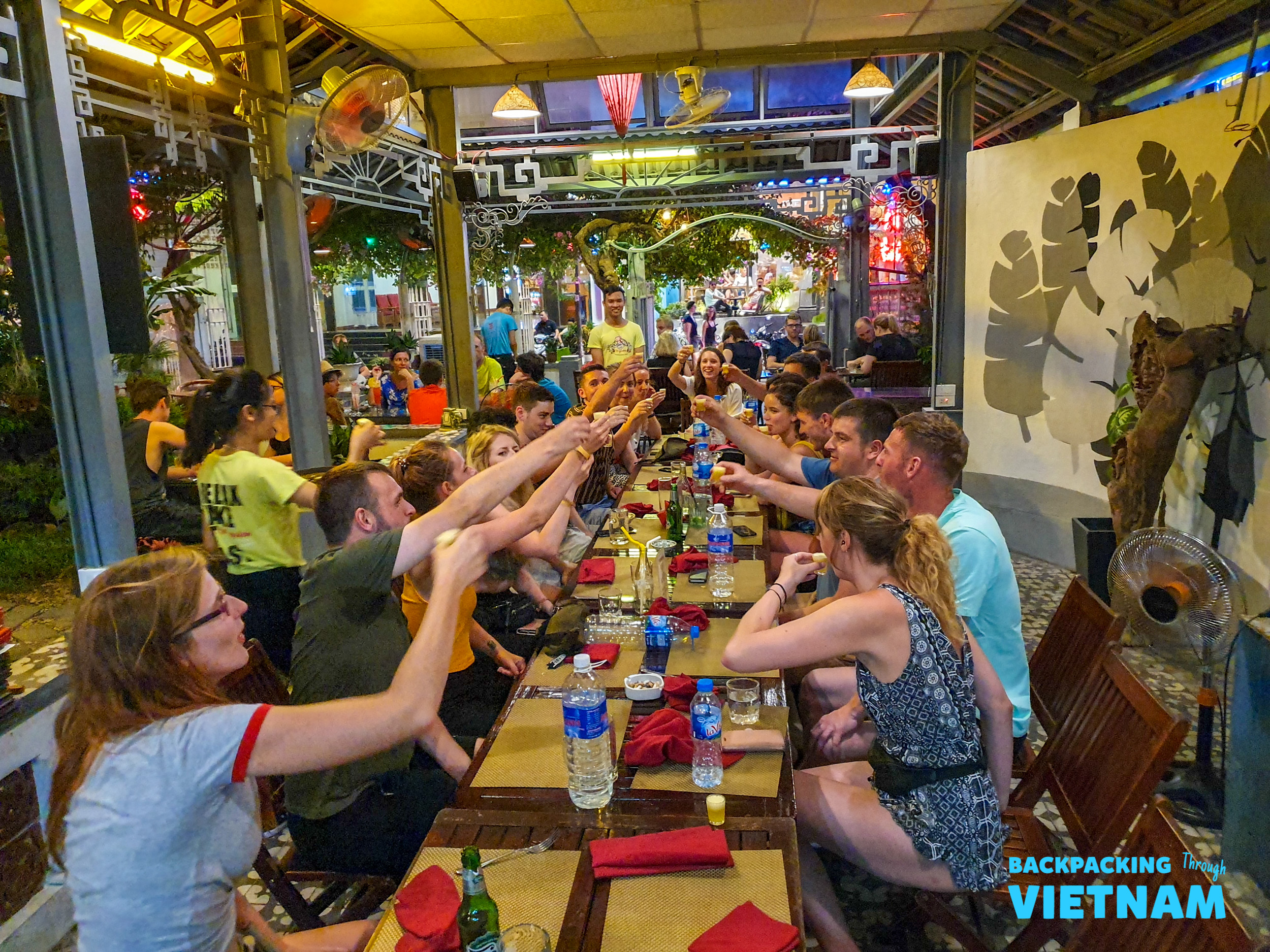 Backpackers toast with drinks at a lively outdoor restaurant in Hue, enjoying the evening.