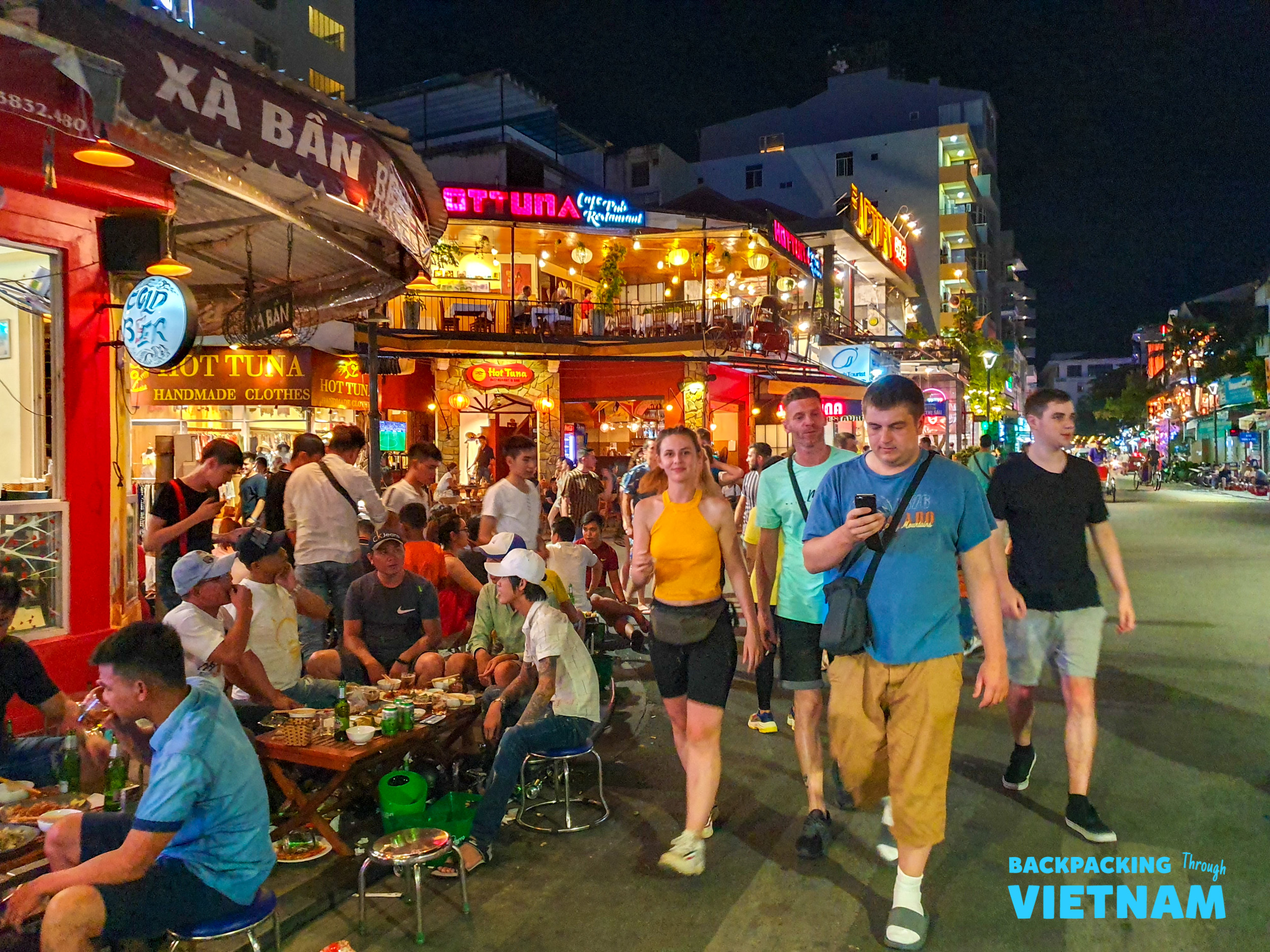 Travelers walk past bustling outdoor restaurants and street food stalls in vibrant Hue at night.