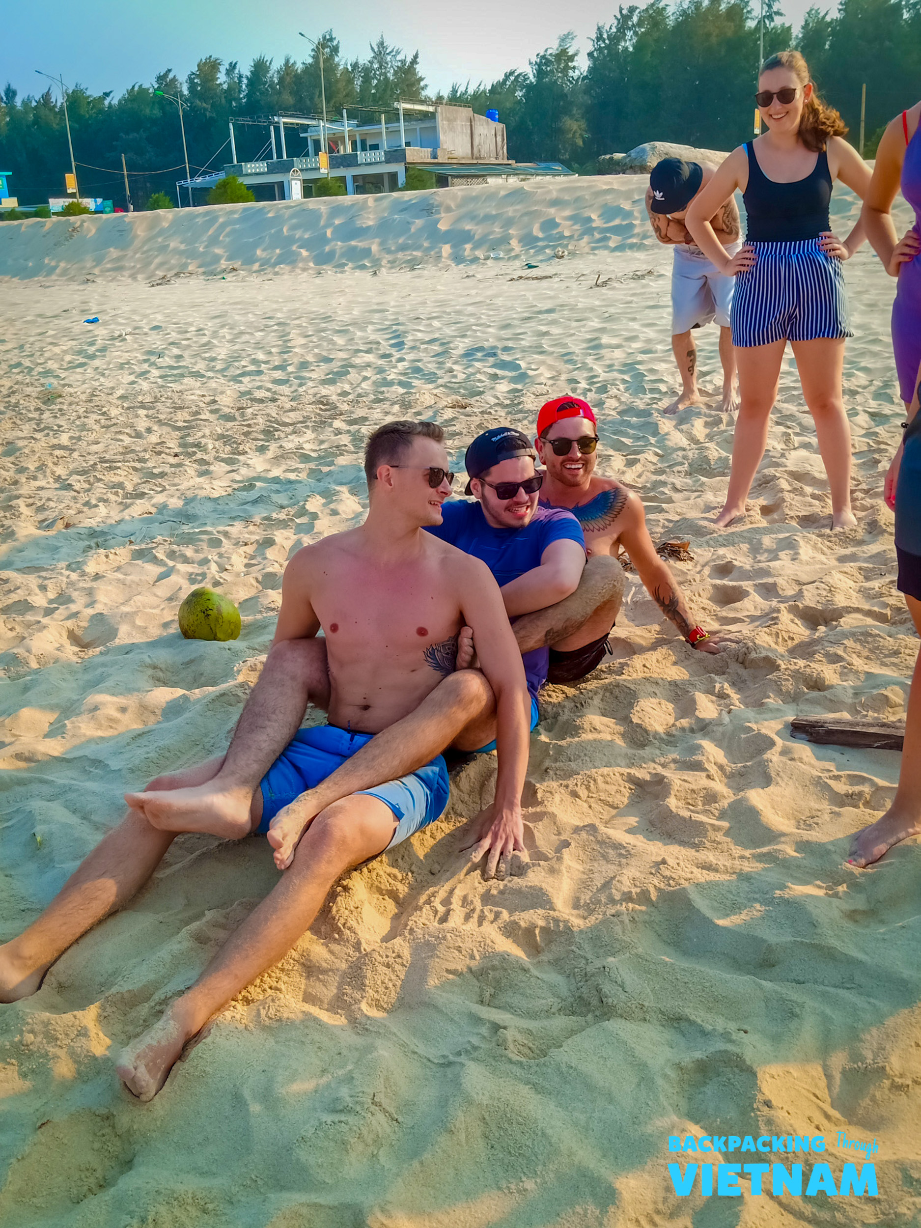Backpackers enjoy a sunny beach day, sitting and laughing on the sand in Vietnam.