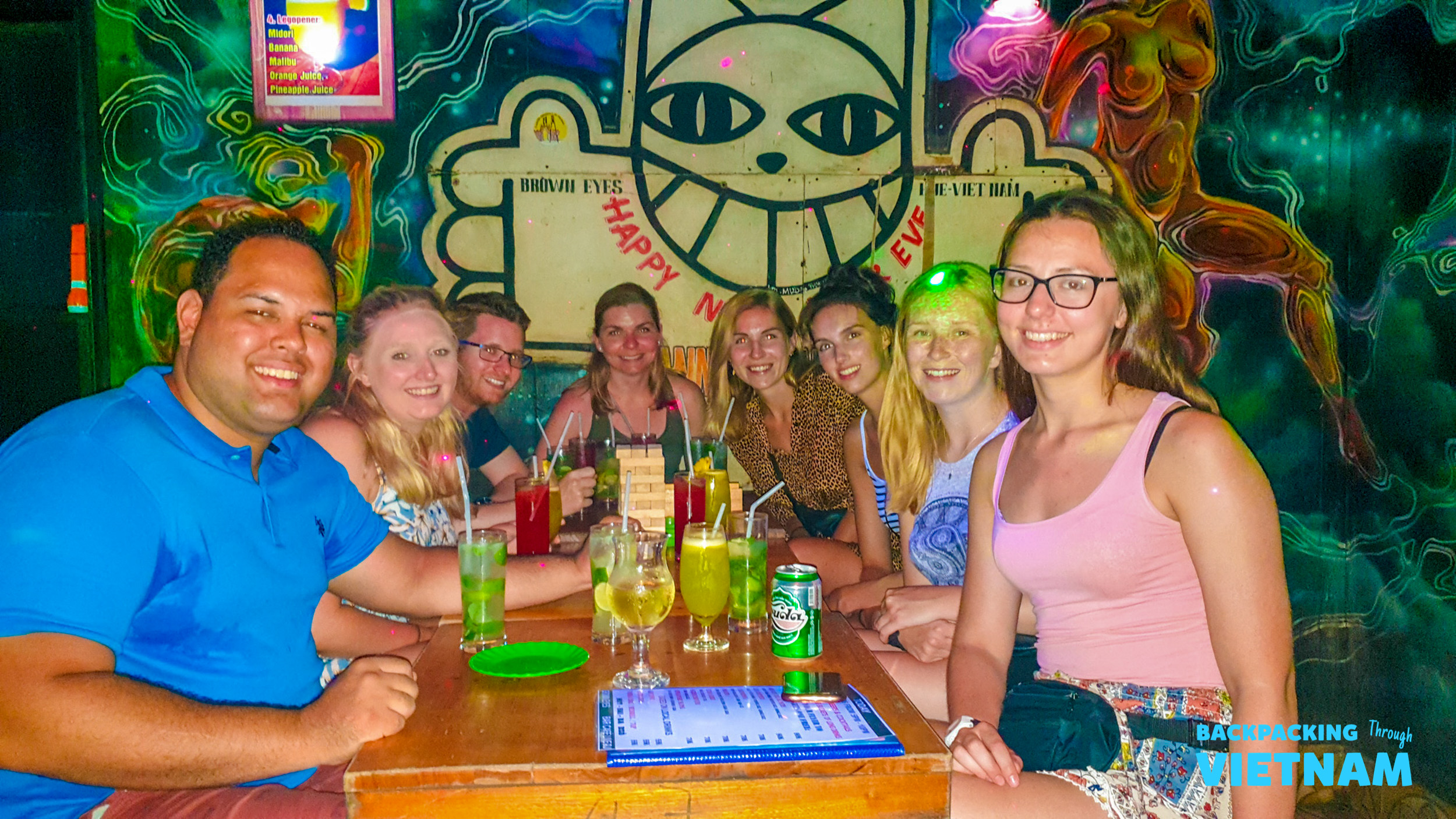 Happy backpackers enjoy colorful drinks at a vibrant bar with neon art in Hue.