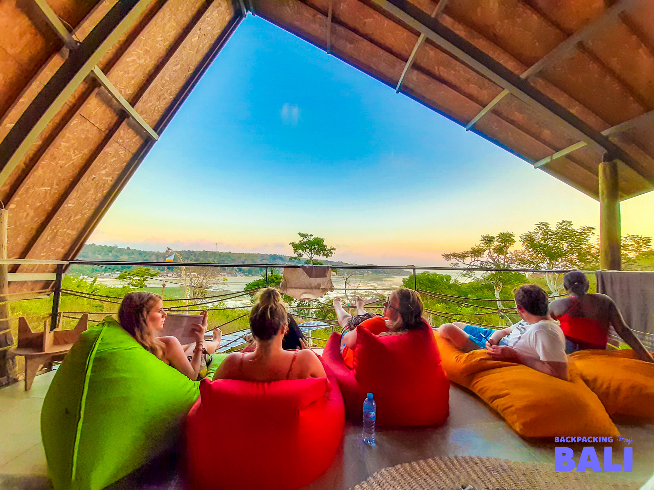 Five friends relax on colorful bean bags on a Bali balcony overlooking a scenic sunset.