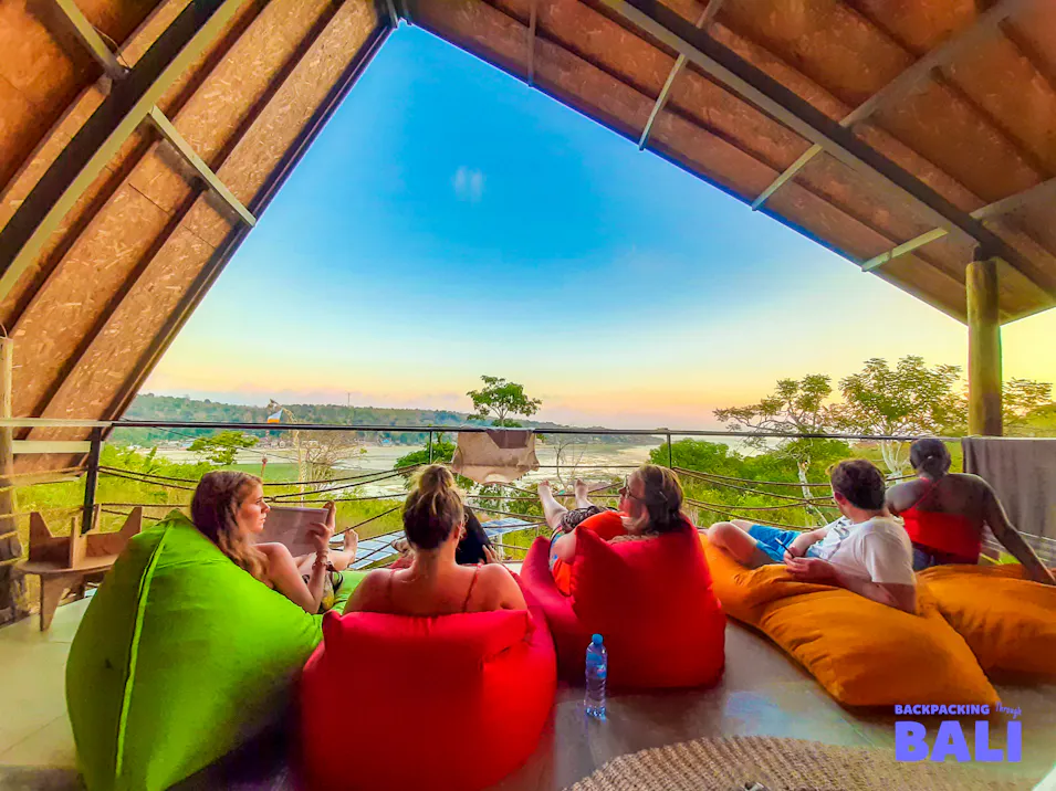 Five friends relax on colorful bean bags on a Bali balcony overlooking a scenic sunset.