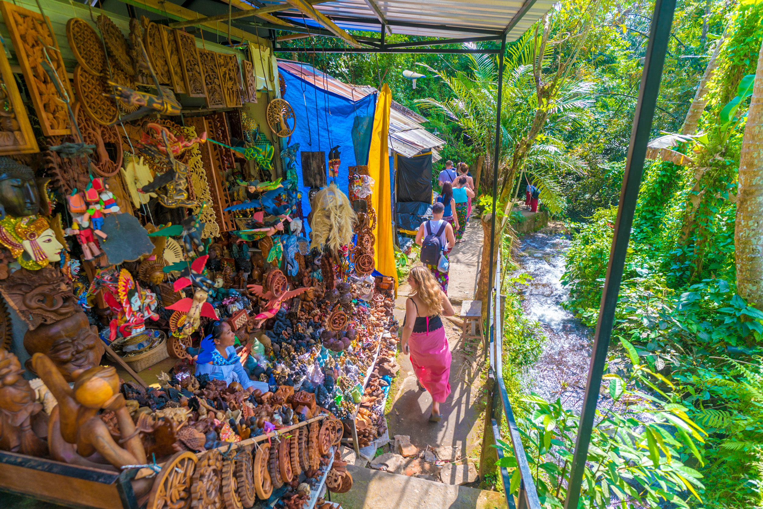 Travelers browse a vibrant outdoor market stall with carved crafts next to a jungle stream in Bali.
