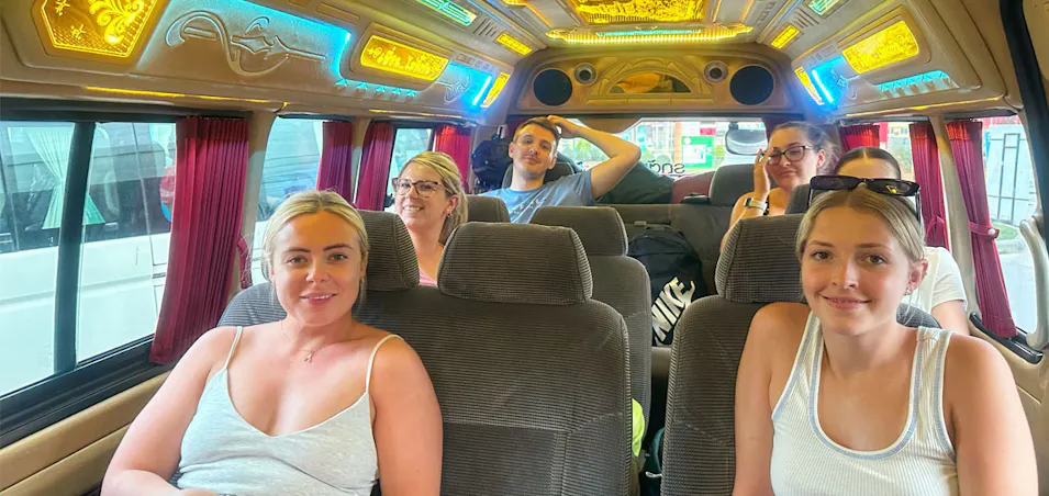 Six happy young travelers smiling inside a colorful, decorated tour van in Thailand.