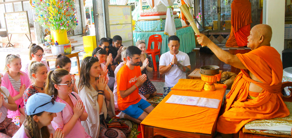 Elderly monk in orange robes blessing a diverse group of people kneeling respectfully in a Thai temple.