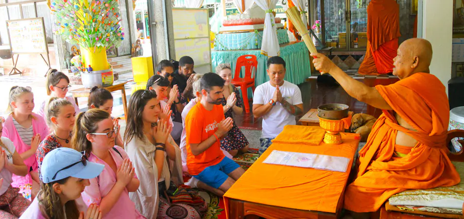 Elderly monk in orange robes blessing a diverse group of people kneeling respectfully in a Thai temple.