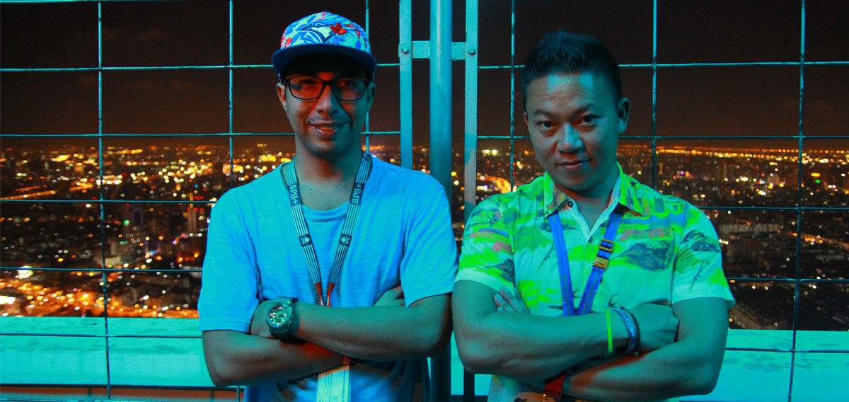 Two men with crossed arms pose on a Bangkok rooftop overlooking a vibrant city at night.