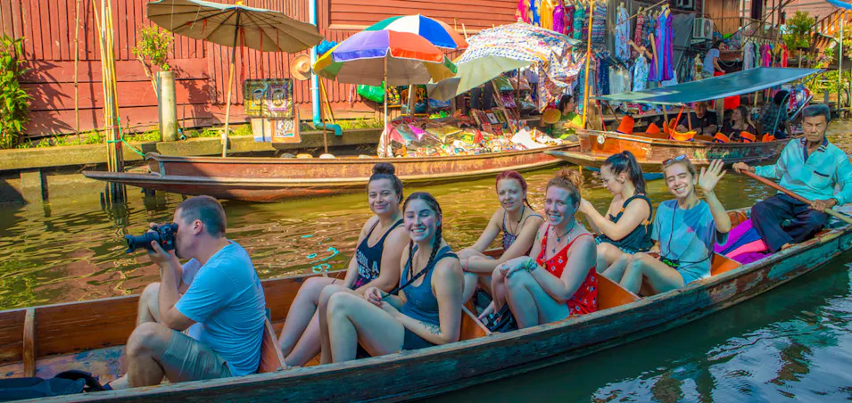 Smiling tourists on a traditional boat exploring a bustling floating market in Thailand with a local guide.