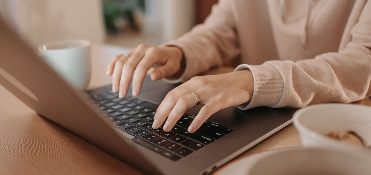 A person's hands typing on a laptop, with a coffee cup and a bowl on a wooden table.