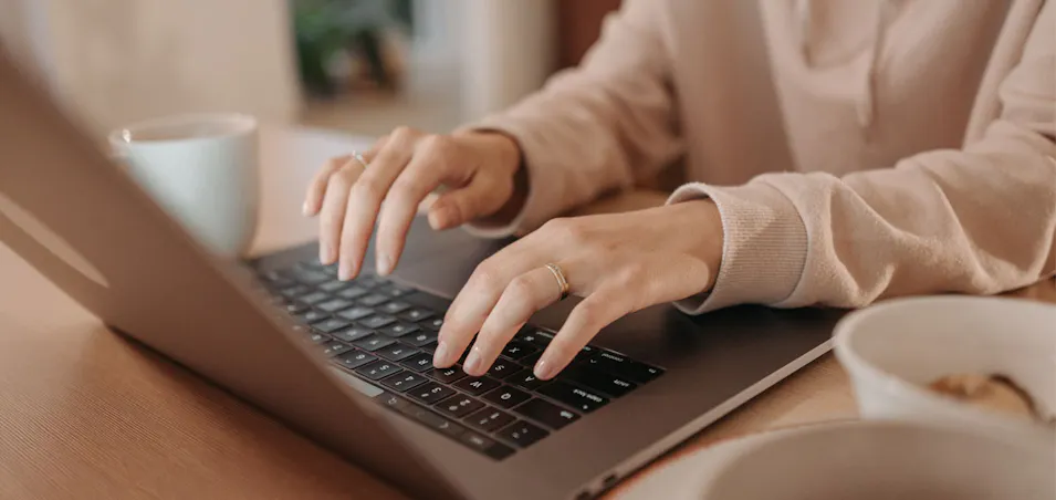 A person's hands typing on a laptop, with a coffee cup and a bowl on a wooden table.