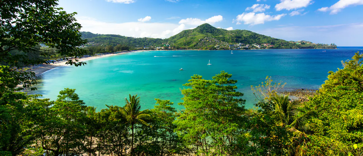 Tropical trees frame a vibrant turquoise bay with a sandy beach and green hills in Phuket.
