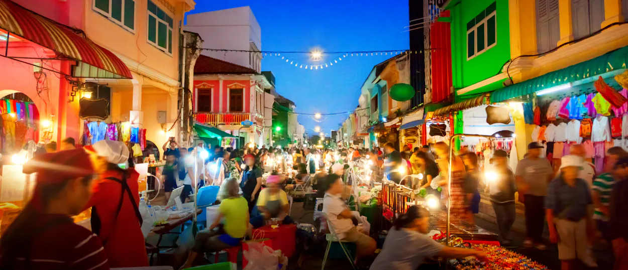 Crowds of people explore a vibrant night market with colorful buildings and bright lights in Phuket.