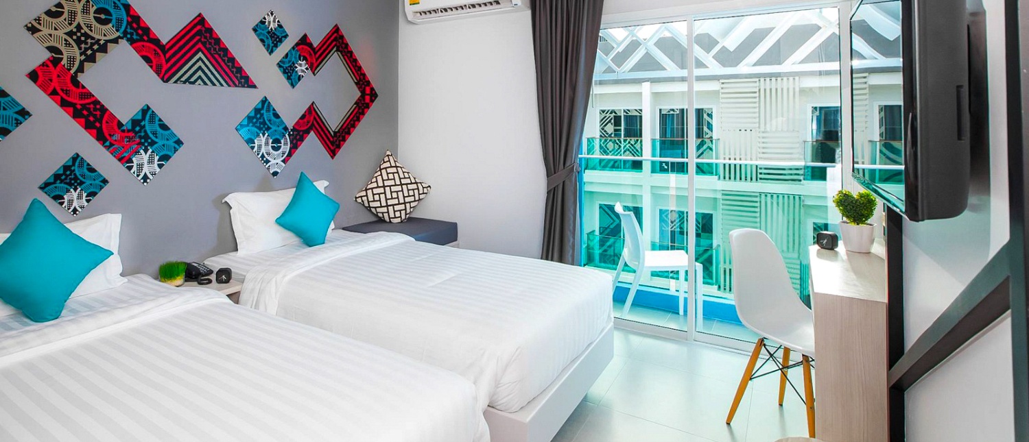 Modern hotel room with two beds, colorful wall art, and a balcony overlooking other buildings.