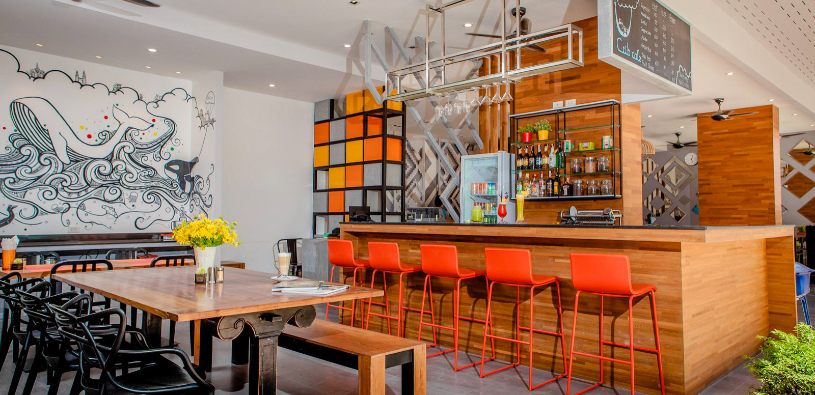 A vibrant, modern bar in Phuket featuring a whale mural, long wooden table, and orange stools.