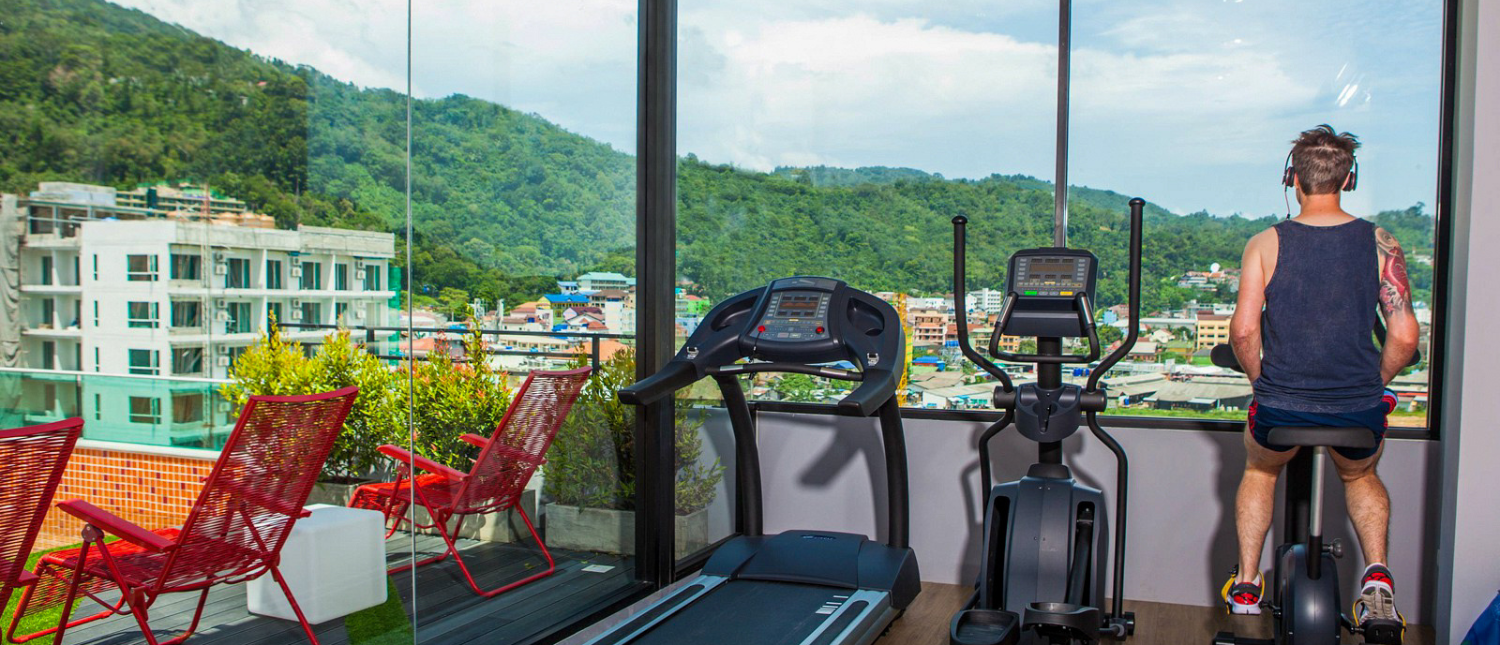 A man exercises on a stationary bike in a gym overlooking green hills and buildings in Phuket.