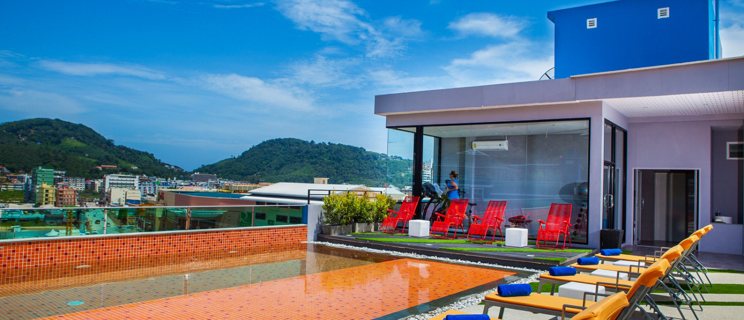 Vibrant rooftop pool with orange loungers and a glass-walled gym overlooking lush green hills in Phuket.
