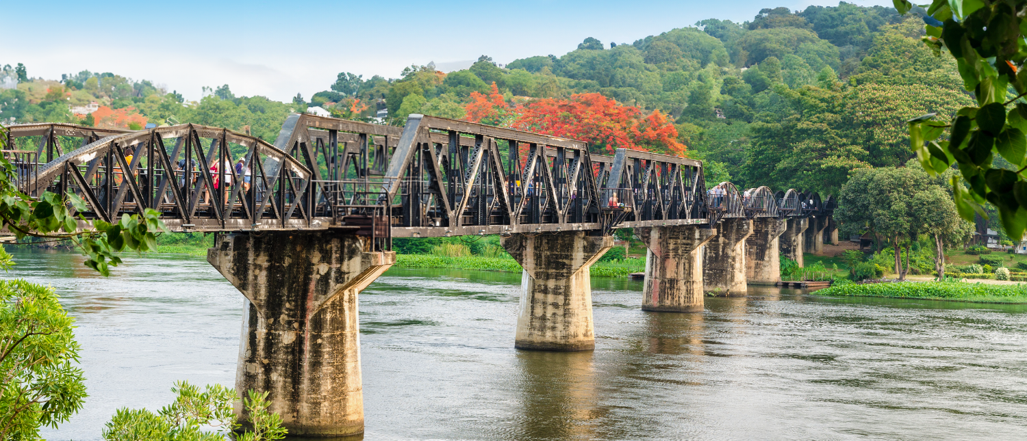 People walk on the historic Bridge on the River Kwai in Kanchanaburi, Thailand, surrounded by lush hills.