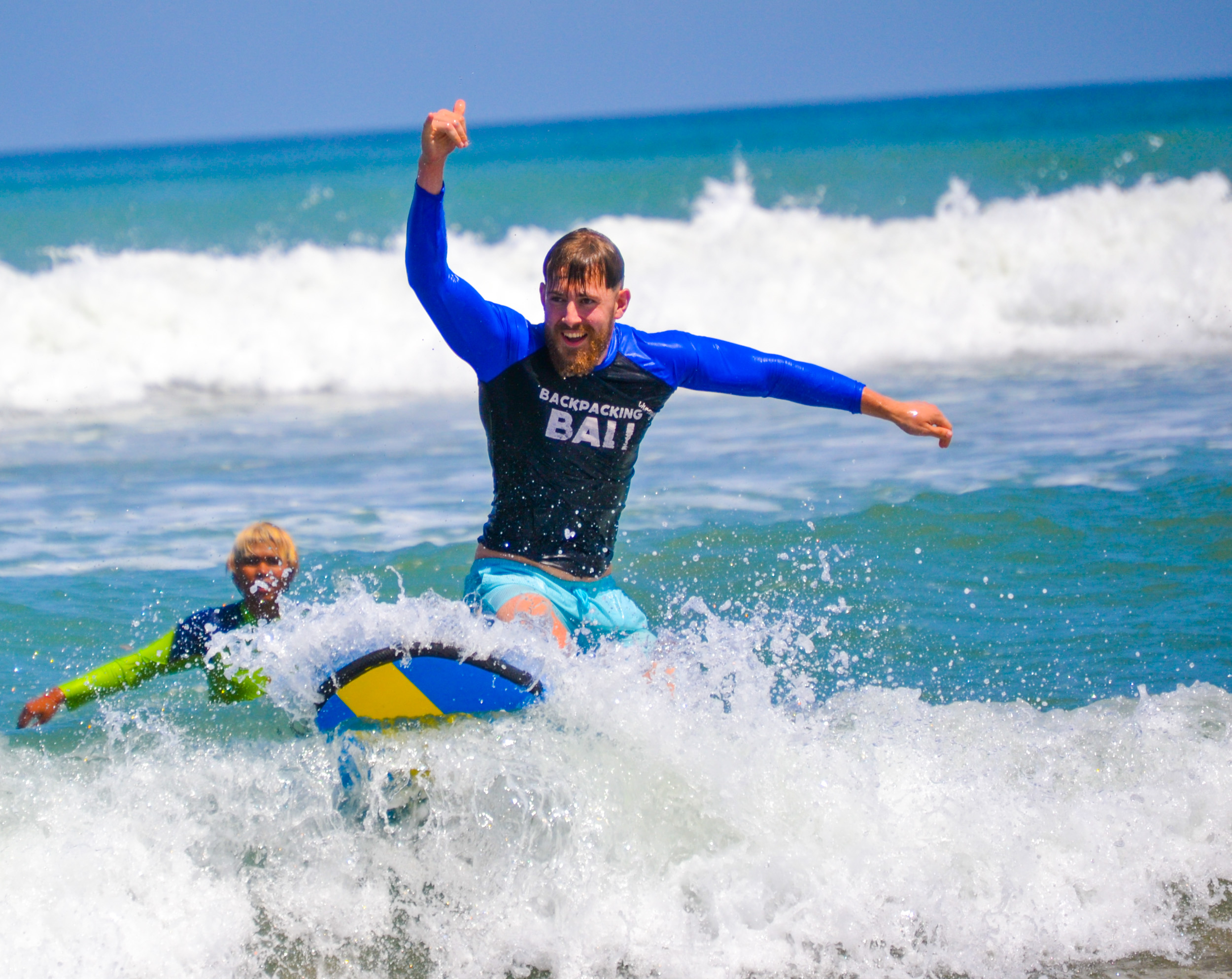 A man joyfully rides a small board on a breaking wave in Bali, cheering with his arm raised.