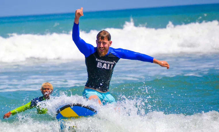 Joyful man in a 'Backpacking Bali' shirt rides a skimboard on a wave, thumbs-up.