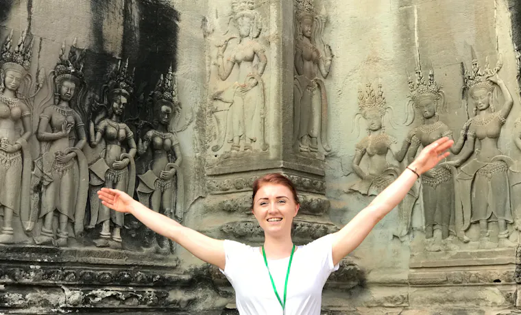 Smiling woman with outstretched arms in front of ancient Angkor Wat temple carvings.