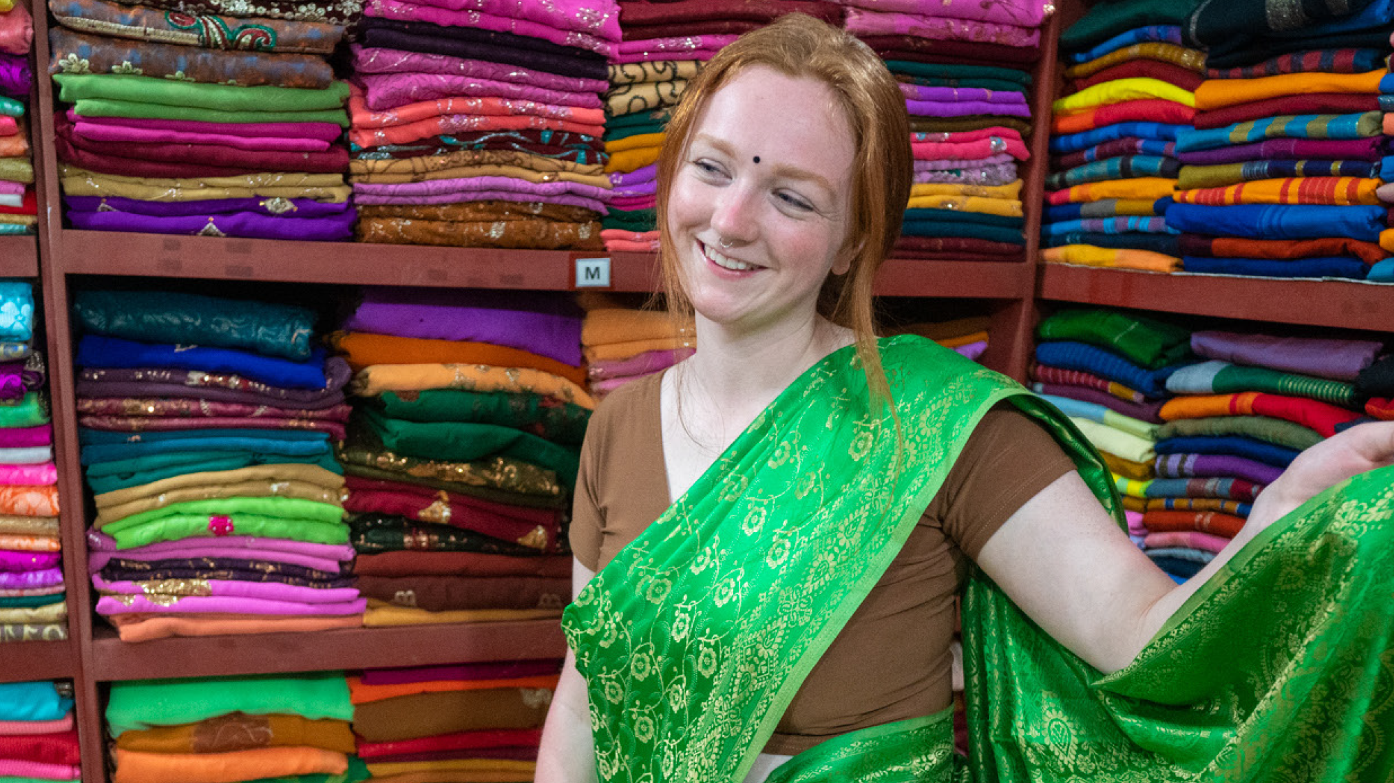 Smiling woman in a green sari among colorful fabric stacks in a Sri Lankan shop.