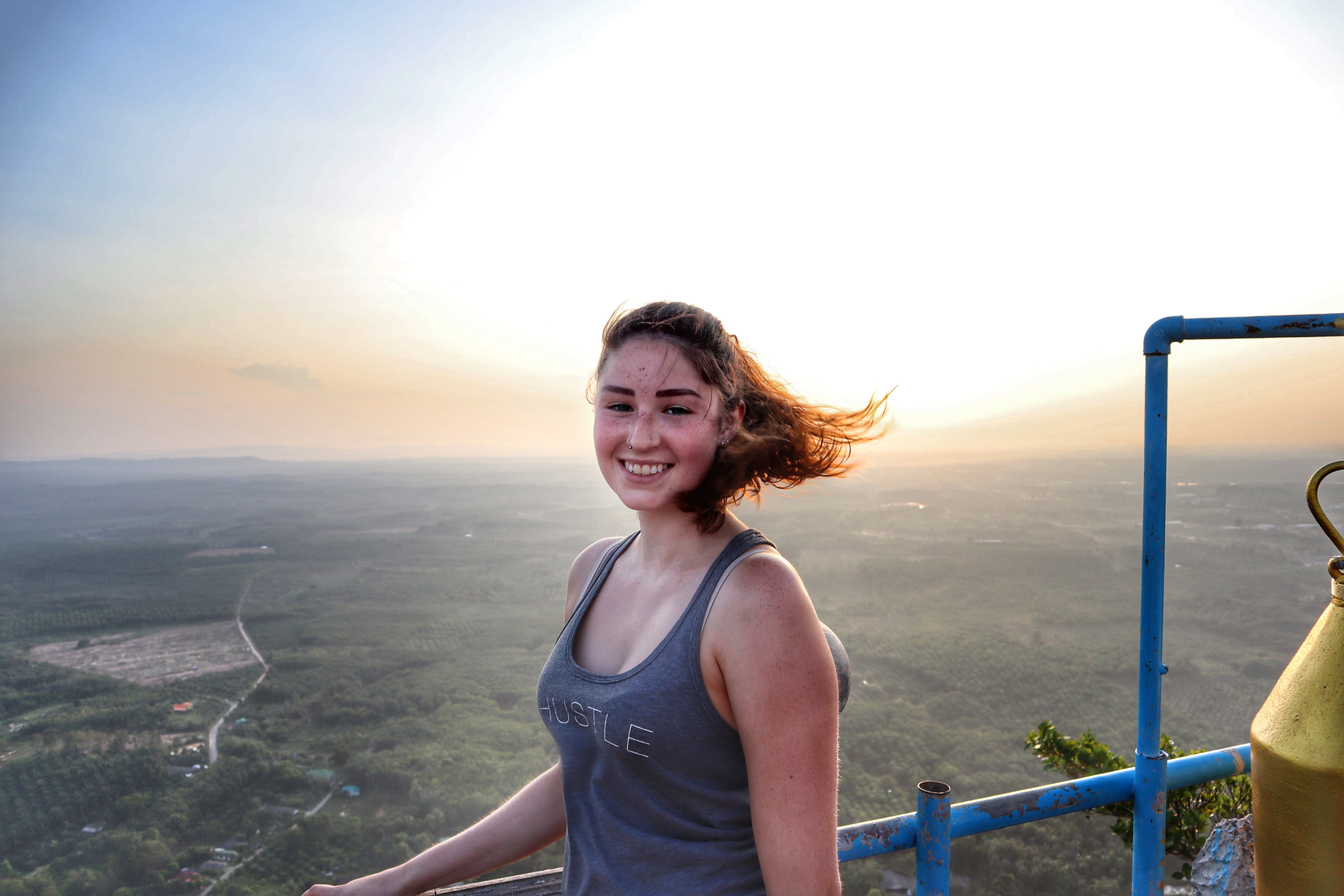 Smiling woman with wind-blown hair on a high viewpoint overlooking a vast Thai landscape at sunset.