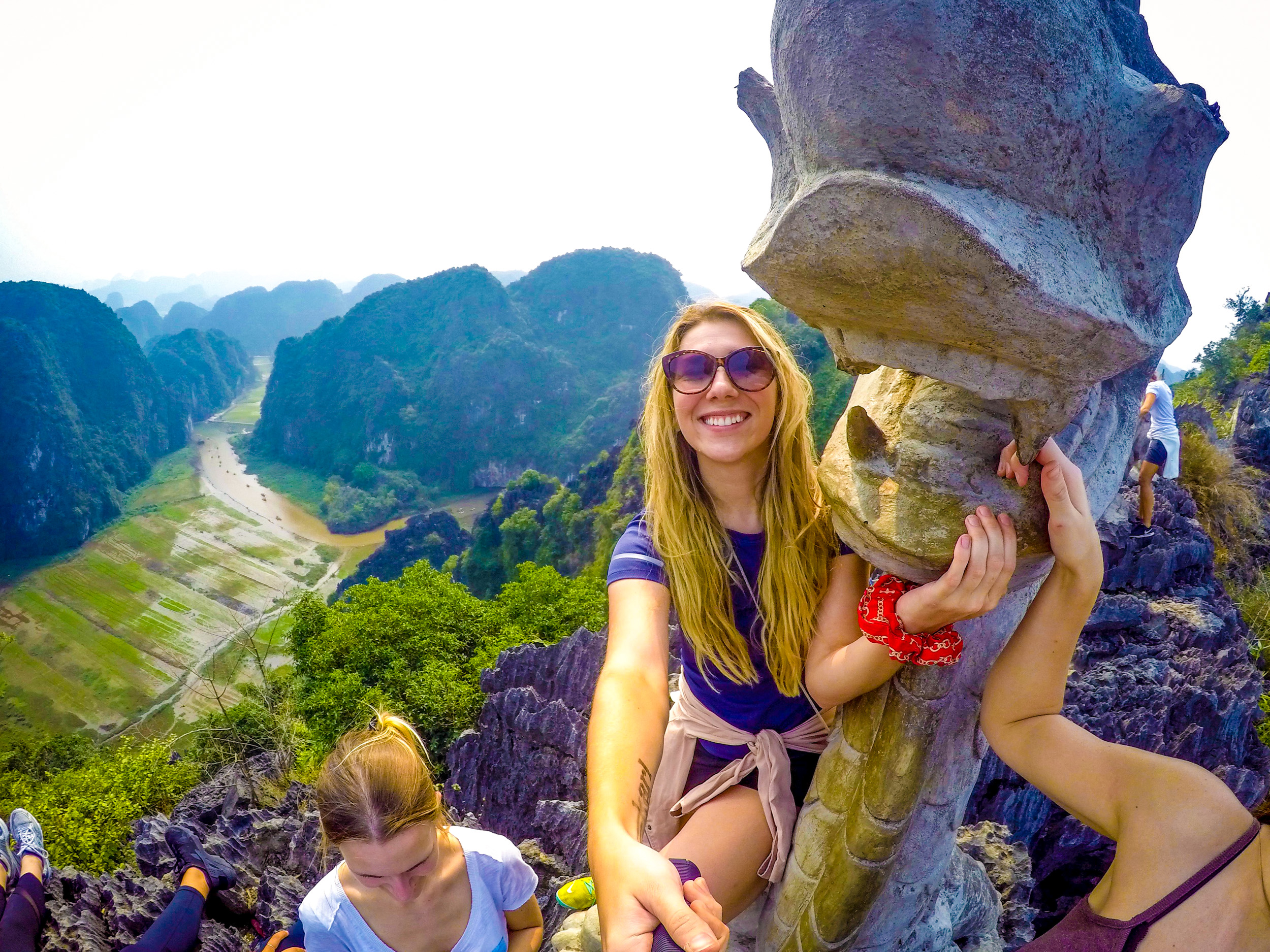 Smiling woman takes a selfie on a rocky peak overlooking a beautiful Vietnamese karst landscape.