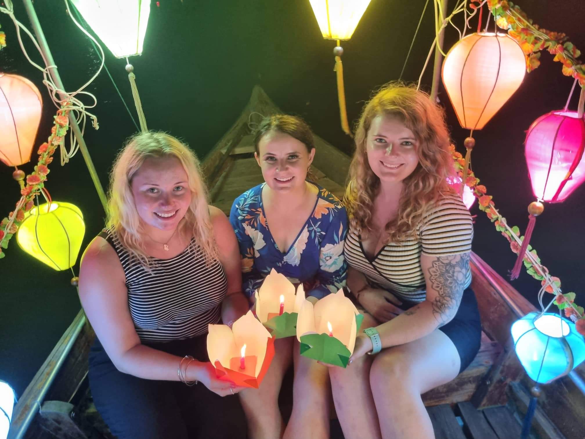 Three happy women in a boat at night, holding candle lanterns amidst many colorful floating lanterns in Vietnam.