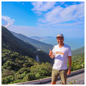 A man smiles and gives a thumbs-up on a mountain road overlooking a beautiful coastline.