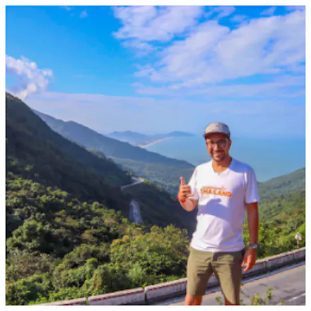 A man smiles and gives a thumbs-up on a mountain road overlooking a beautiful coastline.