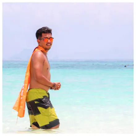 A smiling man wearing sunglasses and swim shorts wades in clear turquoise ocean water.