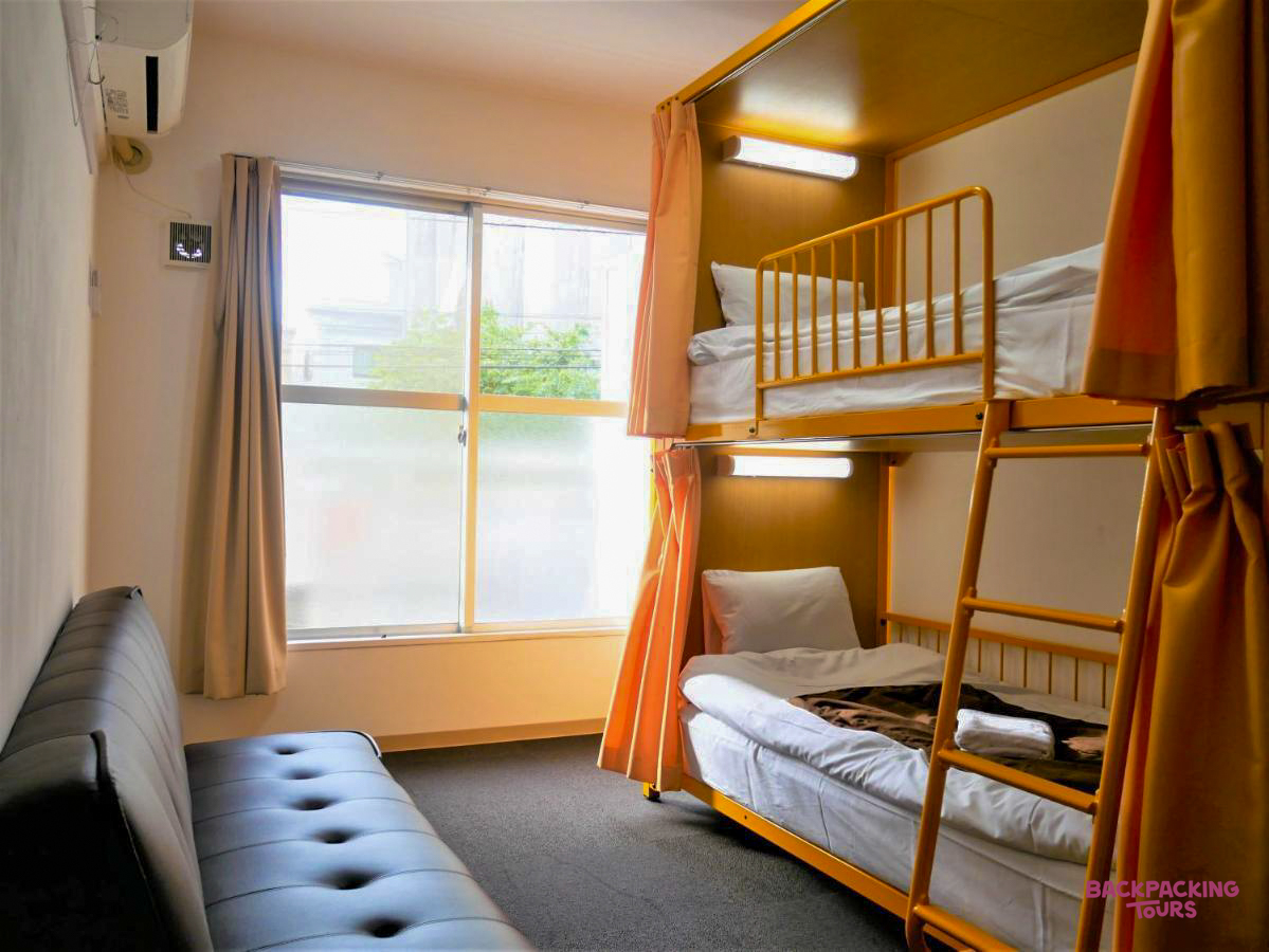 Bright and functional hostel room in Tokyo, featuring a bunk bed with privacy curtains and a sofa.