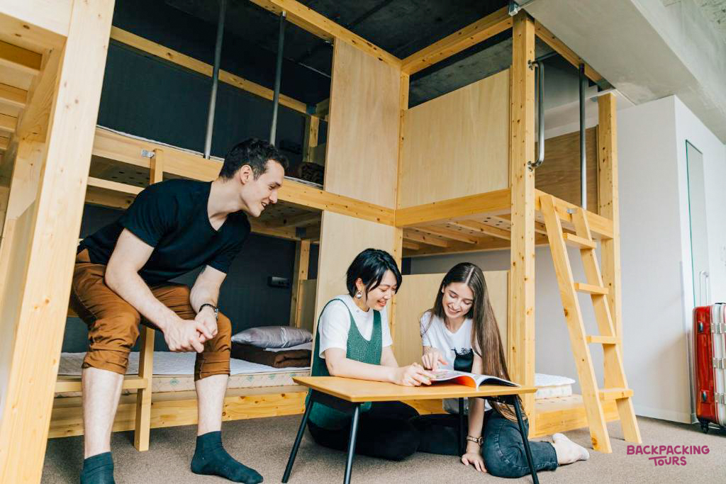 Three happy travelers relaxing and reading in a modern, shared hostel dorm room in Osaka.