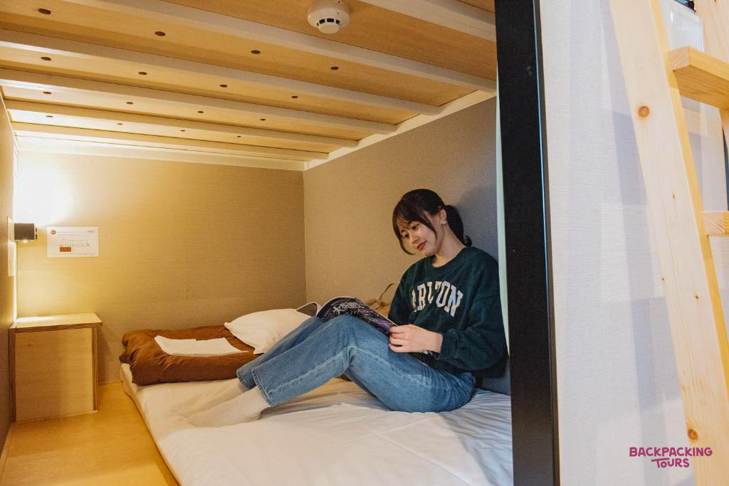 A backpacker relaxes in her cozy capsule bed, reading a book in a hostel in Osaka.