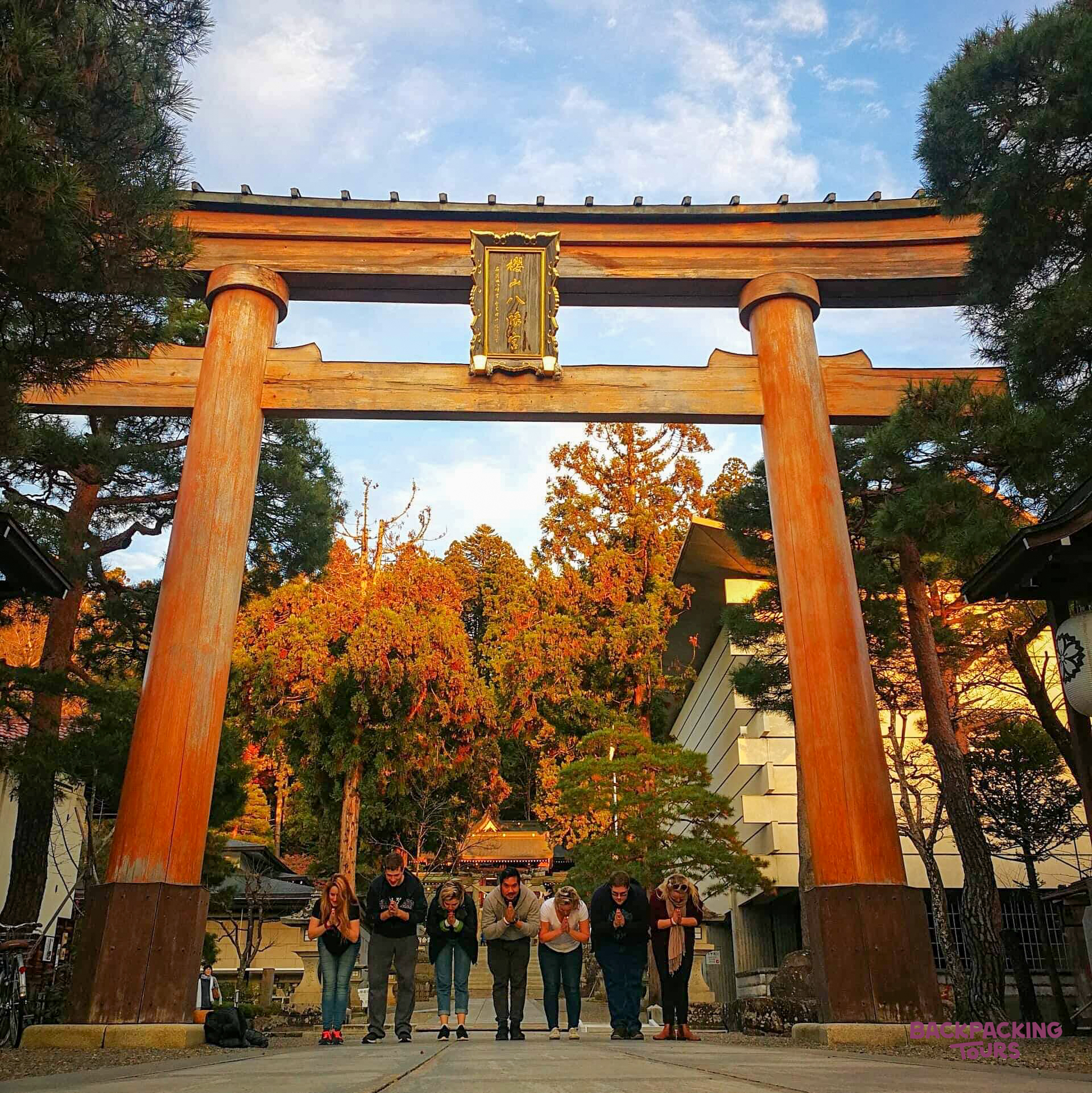 Seven travelers bow respectfully before a vibrant orange torii gate on Miyajima Island.