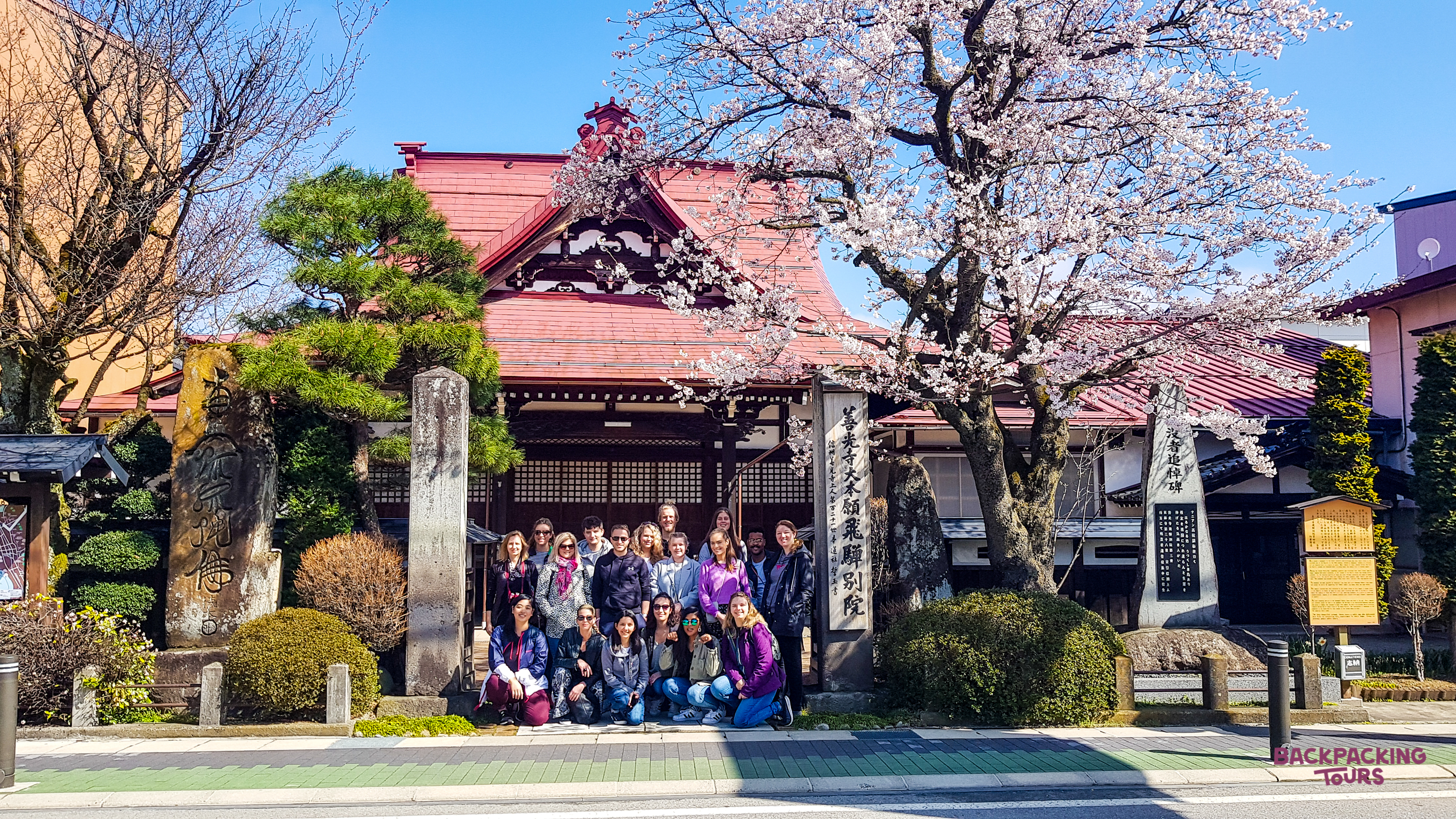 A group of happy backpackers pose in front of a traditional Japanese temple with cherry blossoms in Hiroshima.